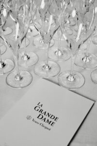 A collection of elegant wine glasses is arranged closely together, reflecting light across their polished surfaces. Below them, a smooth surface lays beneath a sophisticated booklet, exuding an air of refinement. The monochrome theme enhances the chic, formal aesthetic, capturing a sense of anticipation and celebration associated with fine dining or an exclusive event.