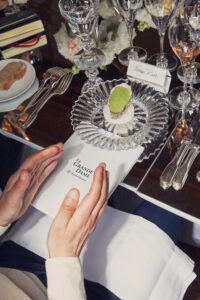 A formal dining setting features an elegantly arranged plate with an oyster on a bed of salt, accompanied by sparkling glassware and a neatly folded napkin. Delicately resting cutlery is positioned nearby. A slice of bread sits on a small plate to the side. Hands hover close to an open booklet, adding a sense of anticipation.