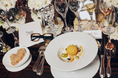 A sophisticated table setting features elegant glassware and white floral arrangements. A beautifully plated dish with a spherical fritter and garnishes sits on a white plate, accompanied by a smaller plate with crusty bread. Two forks and a knife are neatly placed, and a pair of eyeglasses rests nearby. The ambiance suggests a luxurious dining experience.