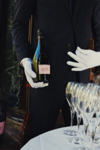 A person wearing a dark suit and white gloves holds a bottle of champagne, preparing to pour it into an array of empty, elegant champagne flutes. The flutes are neatly arranged on a round table with a white tablecloth. The scene exudes an air of sophistication, perhaps set in a formal or high-class event.