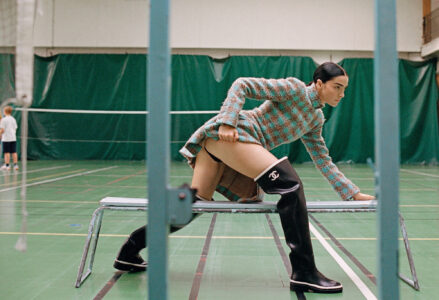 A person in a checkered outfit strikes a dynamic pose on a bench in a gymnasium. The attire includes knee-high black boots and a knee-length jacket. The gymnasium features green walls and a badminton court. In the background, a child in sportswear is engaged in play, adding a sense of motion and activity to the scene.
