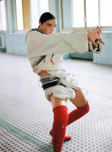 A person strikes a dynamic, expressive pose in an open, brightly-lit space. They wear a layered outfit with a large white jacket and red socks. The floor features a checkered pattern, and tall windows line the walls. The pose exudes energy and movement, with an outstretched arm and bent knee adding to the dramatic effect.