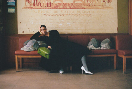 A person sits comfortably on a wooden bench with a red cushion, exuding a relaxed demeanor. They wear a black outfit paired with distinctive white high heels. Beside them, a vibrant green bag adds a pop of color. The backdrop features an artistic, vintage-style mural, creating a warm, inviting atmosphere in the room.