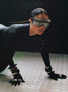 A person in a futuristic outfit poses on all fours on a textured metallic surface. The individual wears dark clothing, large reflective goggles, and has long, wet hair. Their ensemble includes multiple chunky bracelets on both wrists and gloves. The background is dark, enhancing the sleek and avant-garde aesthetic of the composition.