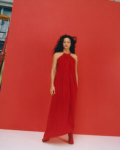 A woman stands gracefully against a vivid red backdrop, wearing a flowing red dress. The scene exudes elegance with her dark, wavy hair cascading naturally down her shoulders. The minimalist setting emphasizes the striking contrast between her attire and the background, creating a harmonious blend of bold color and serene posture, evoking a sense of artistic simplicity.