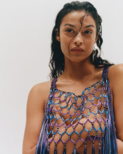 A person with wet, dark hair wears an intricate, netted garment featuring vibrant hues of blue and purple. The accessory showcases a geometric pattern, creating a striking and artistic appearance. A single, abstract silver earring adds a modern touch. The background remains soft and neutral, drawing attention to the poised and confident expression.