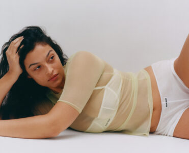 A person with long dark hair reclines thoughtfully. They wear a sheer, pastel yellow top over a structured piece and white bottoms. The background is plain, highlighting the serene expression and relaxed pose. Soft lighting adds a calm and contemplative mood to the scene, focusing on simplicity and elegance. The overall atmosphere is minimalistic and serene.