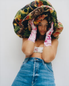 A person stands against a plain backdrop, wearing a floral bikini top and high-waisted jeans. They hold a large, colorful, patterned fabric over their head with both hands, which are covered in pink patterned gloves. The expression is calm and confident, and the overall vibe is artistic and fashion-forward with a focus on vibrant patterns and textures.