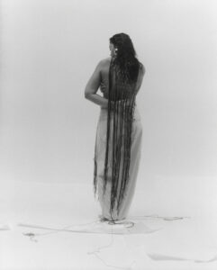A person stands with their back to the viewer, showcasing extremely long, dark hair cascading down their back. They are wearing a long, light-colored garment. The setting is minimalistic, with a plain, light background. The hair extends all the way to the floor, creating a striking visual contrast with the otherwise simple and serene environment.