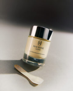 A cylindrical jar featuring a creamy substance is topped with a reflective silver lid, positioned slightly off-center on a neutral surface. Beside it lies a small, flat spatula with a slightly curved shape. The lighting creates soft shadows and highlights, suggesting a serene, minimalist aesthetic. The arrangement implies a focus on skincare or cosmetic indulgence.