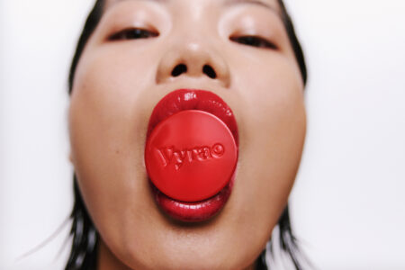 A close-up of a face is shown with lips slightly parted, revealing a glossy red object being held in the mouth. The lips are painted in a matching red shade, creating a bold contrast against the smooth skin. The perspective is from below, highlighting the object's color and texture, while the expression remains enigmatic and striking.