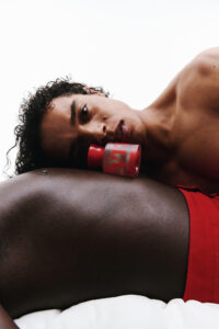 Two individuals are depicted, one resting their head on the other's torso. They are in close proximity, with a small container placed near the person's face. The colors are vivid, focusing on skin tones and red clothing accents. The composition feels intimate and artistic, with a minimalist background enhancing the overall serene and captivating atmosphere.