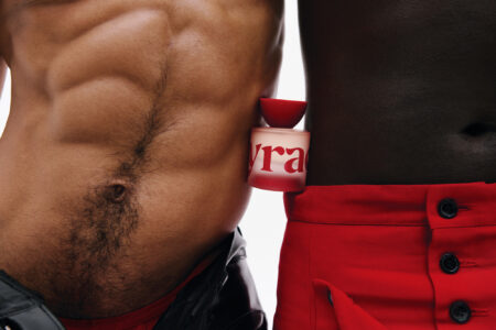 Two individuals stand side by side, showcasing contrasting skin tones. One has well-defined abs and is clad in black clothing, while the other wears vibrant red pants. Between them, a red-capped bottle is nestled, drawing attention to the bold design. The scene emphasizes a harmonious balance of colors and textures, highlighting both fashion and body aesthetics.