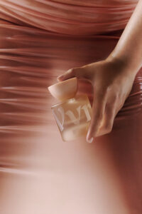 A hand gracefully holds a frosted glass bottle with a softly rounded cap, against a backdrop of smooth, flowing fabric in soft, warm tones. The bottle features bold, elegant designs, complementing the luxurious and sophisticated atmosphere. The composition evokes a sense of elegance and refined simplicity, highlighting the beauty of minimalistic design and gentle colors.