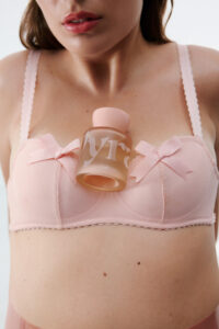A person is featured wearing a light pink bra adorned with delicate bows. Resting on the chest is a frosted pink bottle with a stylish design. The focus is on the detail and texture, capturing the soft fabric and smooth surface of the bottle. The overall color palette is soft and feminine, with emphasis on elegance.