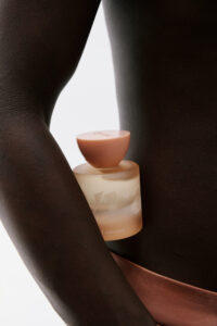 A close-up composition features a matte, frosted bottle with a smooth, rounded cap resting gently against a person's side. The rich, deep skin tone contrasts elegantly with the bottle's muted pastel shades. The arrangement suggests elegance and simplicity, emphasizing the product's form and color against the soft curve of the body, evoking luxury.