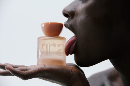 A close-up of a person engaging with a glass bottle, featuring a peach-hued cap. Their hand supports the bottle, with fingers gently curved beneath it. The person’s face is turned to the side, lips slightly parted, and tongue extended towards the bottle. The background is softly illuminated, creating a sense of contrast and focus.