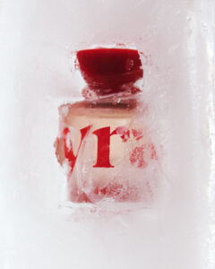 Encased in ice, a transparent bottle with a bold red cap is visible, partially obscured by frost. The scene is dominated by white and red hues, with intriguing textures formed by the ice on the surface. The composition hints at coldness and preservation, contrasting the smooth glass against the rough, frozen surroundings. The overall atmosphere is crisp and refreshing.