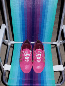Two vibrant pink sneakers with white laces rest on a colorful folding chair. The chair features bold, vertical stripes in shades of blue, purple, and green, adding a playful contrast. The sneakers, positioned neatly side by side, display a stylish design with brown accents. The setting creates a lively and trendy atmosphere, blending fashion with a casual vibe.