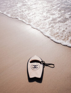 With a serene beach setting, gentle waves softly meet the sandy shore under a bright sky. A sleek and shiny wooden-bodied handboard, accented in black, rests near the water's edge. Its leash lies beside it, casting faint shadows on the sand. The sunlight shimmers on the water, creating a peaceful and relaxed coastal scene.