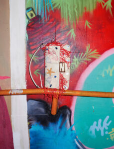 A vividly painted wall features a mix of abstract and whimsical elements. Bright shades of red, green, blue, and pink intertwine with abstract patterns resembling foliage. An orange pipe intersects the scene. A quirky object with starfish and abstract designs, adorned with red string, hangs against the intricate background, contrasting with its playful surrealism.