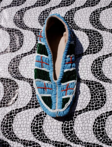 A colorful beaded shoe rests on a black and white patterned surface. The beads on the shoe feature intricate designs with primarily blue, green, white, and hints of red. The background displays a mosaic-like pattern with black arches and white, crackled textures, enhancing the artistic and vibrant contrast between the shoe and the surface beneath it.