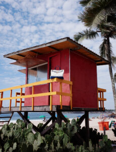 A vibrant scene features a red lifeguard tower with yellow railings perched above a sandy beach. Palm trees sway in the gentle breeze, while people enjoy the sun and sea. A clear blue sky with scattered clouds complements the relaxed atmosphere. Lush green foliage adds a touch of nature, completing the picturesque coastal setting.