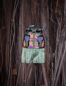 Vibrant floral embroidery in various colors adorns a black surface, with intricate designs framed by light green fringe at the bottom. The colorful pattern includes symmetrical arrangements of blooming flowers and lush leaves. This artistic piece is set against a backdrop of intertwining, textured roots and branches, creating a striking contrast between natural and crafted elements.