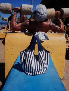 A muscular individual with tattoos lifts large dumbbells outdoors, facing the back. They wear a cap and are positioned against a yellow bench. In the foreground, a striped blue and white bag rests on a blue surface. The setting features palm trees and a clear sky, suggesting a warm, open-air environment suitable for exercising.