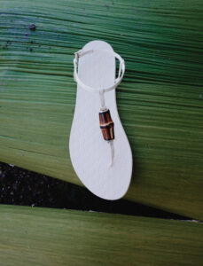 A single white sandal with a brown decorative element lies on a large green leaf. The sandal has a distinct, textured surface and a delicate ankle strap with a buckle. The vibrant, textured green leaf serves as a contrasting background, enhancing the simplicity and elegance of the footwear's design. Shadows and natural light play across both objects.