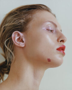 A profile view highlights elegant features and smooth skin, accentuated by vibrant red lips and a soft, glossy eyelid. Light catches the high cheekbone, creating a luminous effect. Hair is styled loosely away from the face, while a small, distinctive birthmark rests near the jawline. Subtle makeup and flattering angles give an artistic, polished appearance.