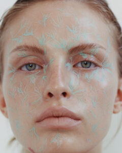 A close-up portrait showcases a person's serene expression with intricate, pastel blue botanical patterns across their face. The individual has soft, natural lips and light eyes that gaze directly ahead. The skin has a dewy, luminous quality, and the hair is styled back. The artistic design creates a captivating fusion of natural beauty and creative expression.