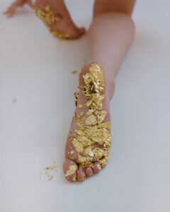 A foot is gracefully extended, partially covered in delicate, metallic gold leaf. The leg is resting on a smooth, white surface, creating a striking contrast between the skin, gold accents, and the background. The scene evokes a sense of luxury and artistry, highlighted by the intricate, scattered patterns of the golden flakes adorning the foot.