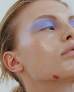 A person with fair skin is highlighted, featuring striking blue eyeshadow adorning their eyelids. Their gaze is directed sideways, with attention to the high cheekbones and subtle lip color. A small, red birthmark is visible near the jawline. The hair is lightly tousled, and the overall ambiance captures a blend of natural beauty with elegant makeup artistry.