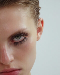 A close-up of a person's face focusing on one eye, showcasing long eyelashes and light-colored eye. The skin has a dewy appearance with defined facial features. Hair is slicked back, revealing an ear. The expression is intense and captivating, highlighted by well-shaped eyebrows and natural makeup tones, creating a minimalist and striking look.