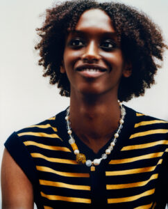 A person smiles softly with curly hair framing their face. They wear a sleeveless black top with bold yellow horizontal stripes. A distinctive necklace featuring a variety of beads, including pearls and an intricate yellow piece, complements the outfit. The background is a simple, smooth gradient with natural lighting, highlighting the subject's expression and attire.