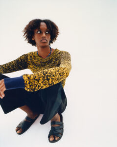 A person with curly hair sits on the ground, looking to the side. They wear a yellow and black patterned sweater, dark wide-legged trousers, and black sandals. A blue necklace adds a pop of color to their outfit. The background is plain and light, emphasizing the relaxed and thoughtful expression on their face.