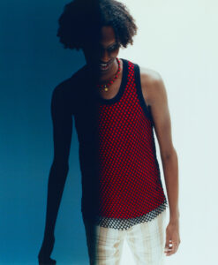 A person stands against a gradient backdrop, blending from blue to light. They're wearing a red and black netted sleeveless top over a red garment and plaid pants. A beaded necklace adds a pop of color. The lighting creates a shadow across half their face, enhancing the contrast between the clothing and the background.