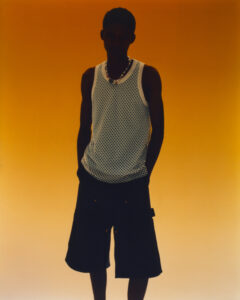 A silhouette of an individual stands against a vibrant orange backdrop, wearing a white sleeveless mesh top and dark shorts. The lighting casts strong shadows, obscuring facial features while accentuating the clothing's texture. A necklace adds detail to the otherwise minimalistic and bold visual composition, creating a striking contrast against the warm, bold background.