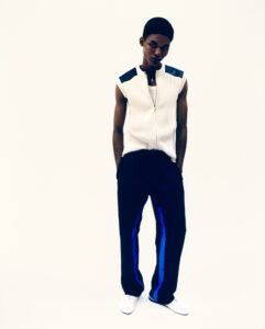 A person stands confidently against a plain backdrop, exuding a sense of modern style. They're wearing a sleeveless, ribbed white top with dark shoulder accents, paired with loose-fitting dark trousers featuring striking blue stripes along the sides. Completing the look are sleek white shoes. The overall impression is one of casual elegance and contemporary fashion.