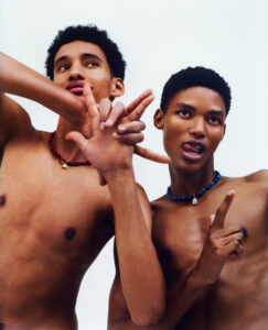 Two individuals pose against a plain background, both wearing beaded necklaces—one in red with a gold pendant and the other in blue with a silver pendant. Their expressions are playful and dynamic, featuring gestures as they interact with the camera. The composition highlights their lively energy and the minimalist aesthetic emphasizes their accessories and expressions.