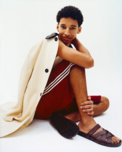 A person sits comfortably with a relaxed posture, one hand resting on a bent knee. They wear a stylish outfit that includes a sleeveless top beneath a draped coat and red shorts with white stripes. Their feet are adorned with dark brown woven sandals. The overall ambiance is casual and modern with a light, minimalistic background.