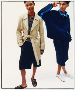 Two individuals stand against a plain backdrop. The person on the left wears a light trench coat over a patterned sweater, paired with dark shorts and sandals. The other sports a dark athletic jacket with blue accents, matching shorts, and white sneakers. Both appear relaxed, exhibiting an urban style with attention to casual yet stylish elements.