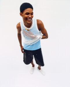 A person joyfully smiles, dressed in a white mesh sleeveless top over a light blue shirt. They wear dark shorts and white sneakers, accessorized with a colorful beaded necklace. The background is plain and soft, emphasizing the casual, vibrant feel of their outfit and expression. The overall mood is playful and carefree.