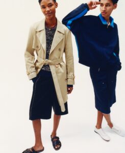 Two individuals stand against a light background. One wears a beige trench coat over a textured sweater with dark shorts and sandals. The other sports a navy zip-up jacket with bright blue accents, dark shorts, and white sneakers. Both appear relaxed and stylish, exuding a modern, youthful vibe. Their poses convey confidence and a casual sense of fashion.