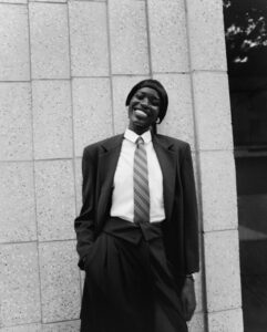 A person with a joyful expression is dressed in formal attire, featuring a dark suit and a striped tie. They are leaning casually against a textured, tiled wall. Their hands are tucked into their pockets, and the background has a distinct speckled pattern. The setting conveys a relaxed yet sophisticated urban vibe, emphasizing confidence and style.