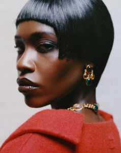 A person gazes sideways, showcasing sleek, short black hair with blunt bangs. They wear a bold red textured garment. Elegantly adorned, gold jewelry featuring colorful gemstones accents the ensemble. The lighting emphasizes their smooth complexion and understated eye makeup. The overall mood conveys a sense of sophistication and poise, blending modern style with classic elegance.