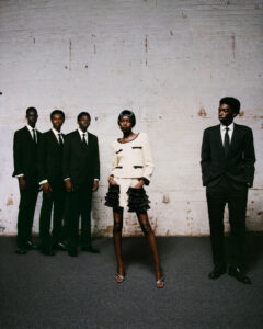 Five individuals stand against a textured, light-colored brick wall. Four wear sharply tailored black suits, exuding elegance and formality, while a central figure stands out in a cream-colored outfit with black accents and ruffled details, adding contrast and intrigue. The setting is minimalistic, with a subtle carpeted floor, enhancing the overall stylish and sophisticated ambiance.