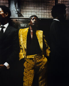 Three individuals are in a dimly lit, industrial setting. The central figure wears a striking yellow patterned suit with hands in pockets, exuding confidence. On either side, two others are dressed in dark suits, adding contrast. A metal grid background and warm, moody lighting emphasize the atmosphere, blending elegance with an urban vibe.