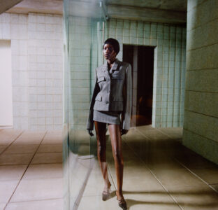 A person stands confidently in a structured, tailored gray suit with a mini skirt, exuding elegance and modern style. The background features an architectural space with pale tiled walls and a reflective glass surface. The individual wears a sleek hat, and their striking posture is emphasized by soft lighting, which casts subtle shadows on the floor.